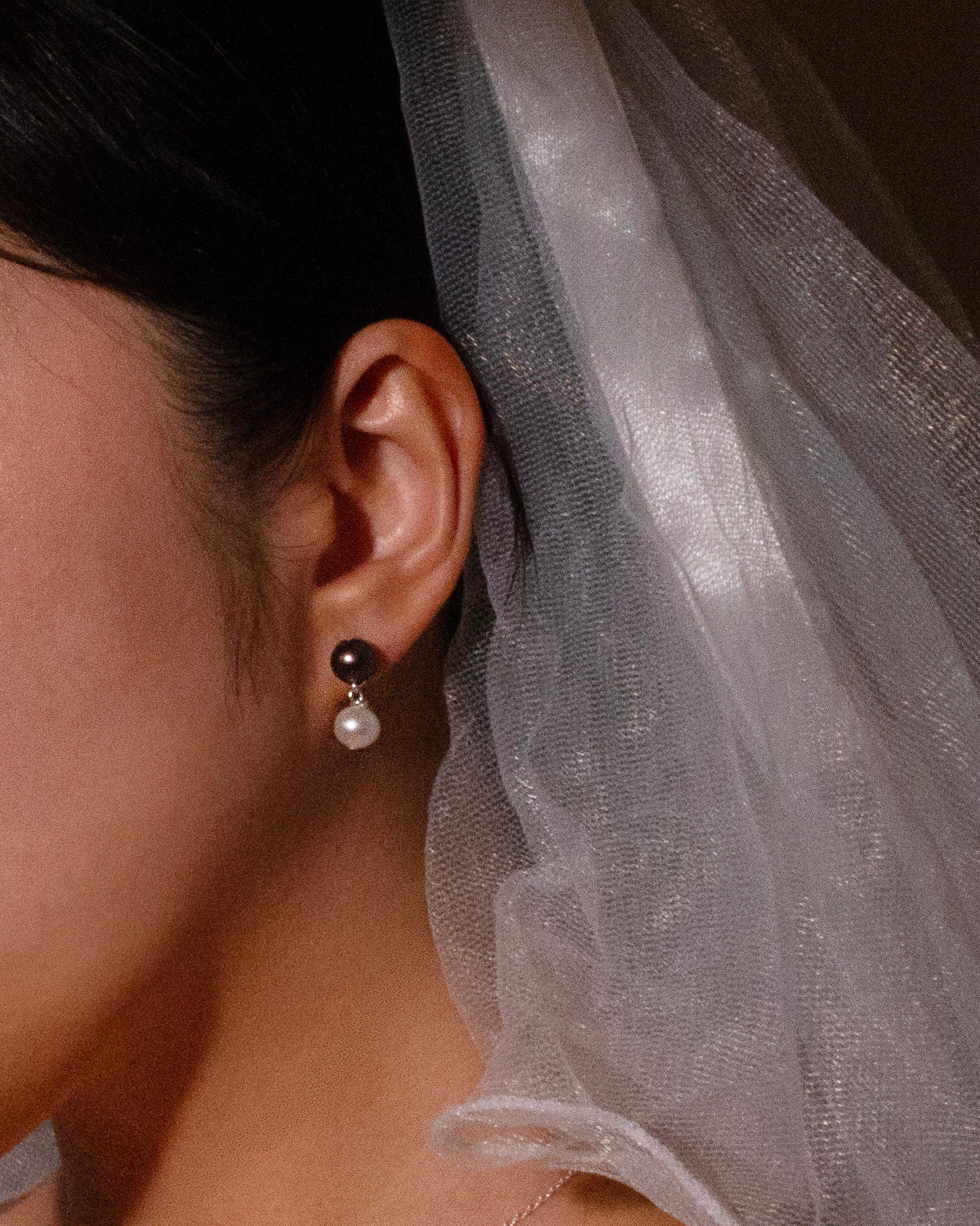 Close-up of a person wearing a black and pearl earring with a veil in the background