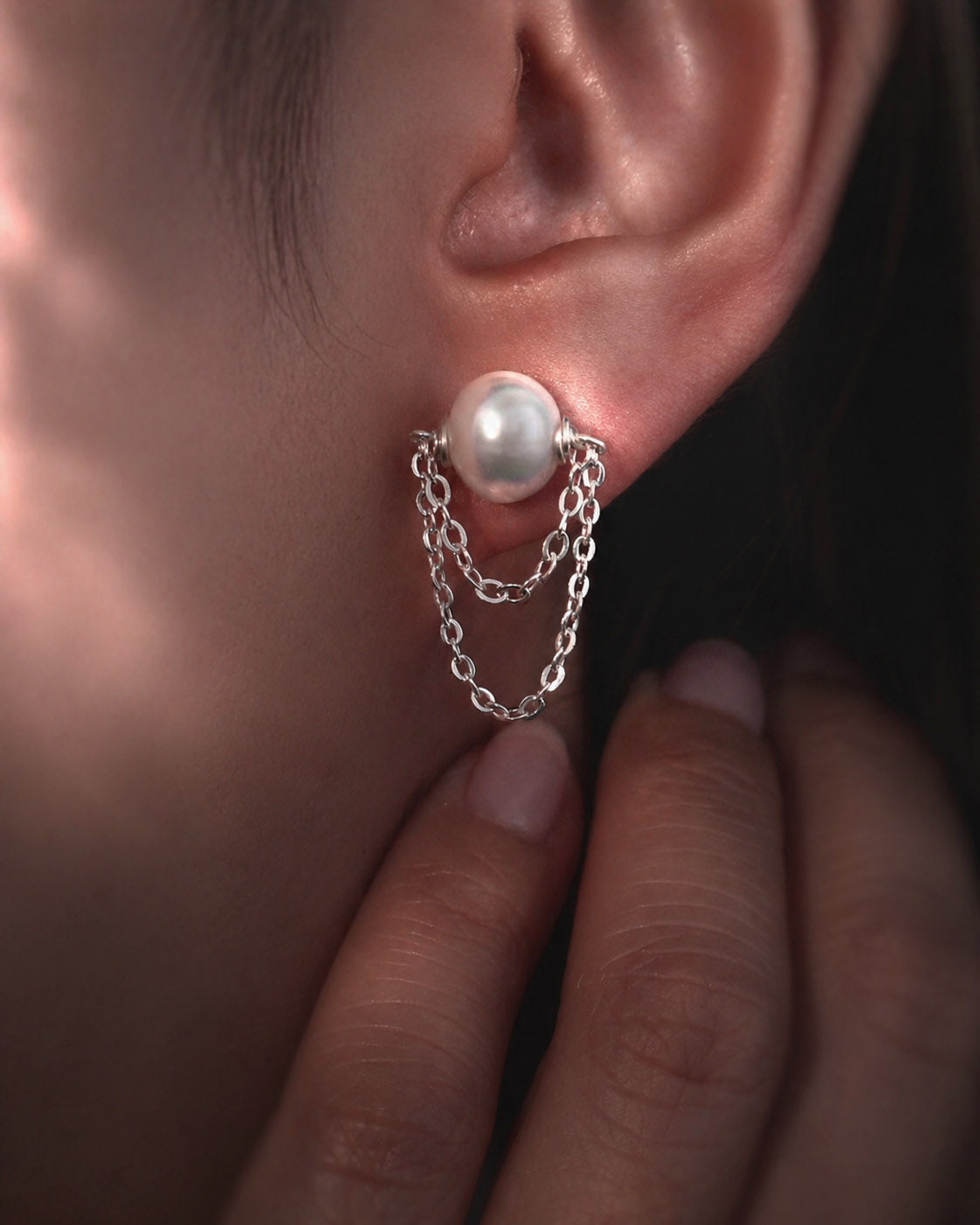 Pearl earring with chain detail worn by a person, close-up of ear and hand.